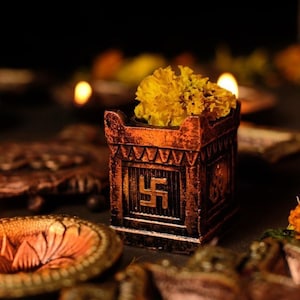 May include: A small, ornate, wooden box with a swastika symbol on the front. The box is filled with yellow marigold flowers. The box is surrounded by other decorative items, including small, round, metal bowls.