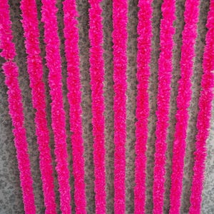 May include: Vertical rows of vibrant fuchsia-colored, textured garlands hang against a speckled gray background. The garlands appear to be made of soft, ruffled material, creating a visually appealing, decorative display.