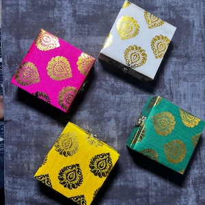 May include: Four small, square boxes in vibrant colors: hot pink, white, yellow, and teal. Each box is adorned with a gold paisley pattern. The boxes are closed and appear to be made of fabric.