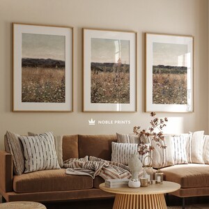May include: A brown sectional sofa with a wooden coffee table and a woven ottoman in front of it. The sofa has a variety of throw pillows and a blanket. Three framed landscape prints hang on the wall behind the sofa. The text "NOBLE PRINTS" is printed below the prints.