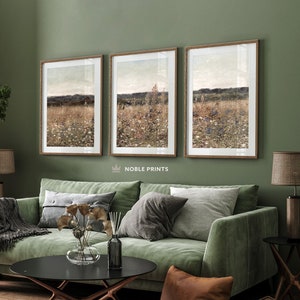 May include: Three framed landscape paintings of a field of wildflowers with a distant horizon. The paintings are hung on a sage green wall above a green velvet sofa with brown throw pillows.