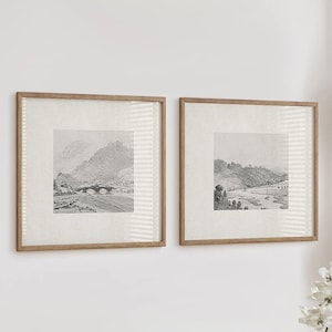 May include: Two framed black and white landscape photographs hanging on a white wall. The photos are in wooden frames with a light wood finish. The photos are of a mountain range and a river.