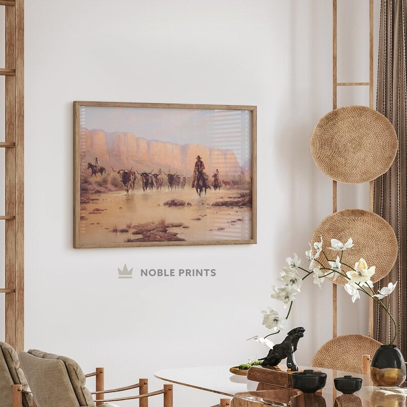 May include: A framed print of a vintage Western scene depicting a group of cowboys on horseback driving a herd of cattle through a shallow river. The scene is set against a backdrop of rugged cliffs and a hazy sky.