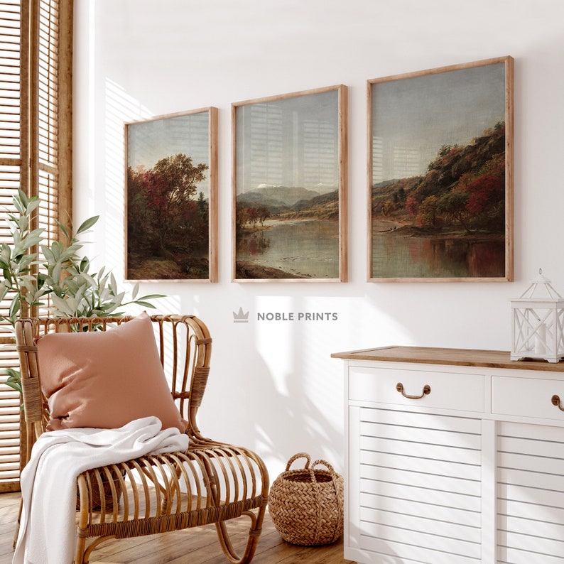May include: Three framed landscape paintings with a rustic aesthetic. The paintings depict a river scene with trees and mountains in the background. The paintings are hung on a white wall in a living room setting.