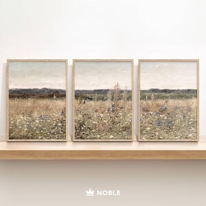 May include: Three framed prints of a field of wildflowers in shades of blue, purple, and white. The field is set against a hazy, light blue sky and a distant line of trees. The frames are a light wood color.