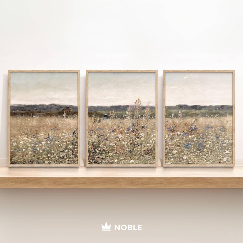 May include: Three framed prints of a field of wildflowers in shades of blue, purple, and white. The field is set against a hazy, light blue sky and a distant line of trees. The frames are a light wood color.