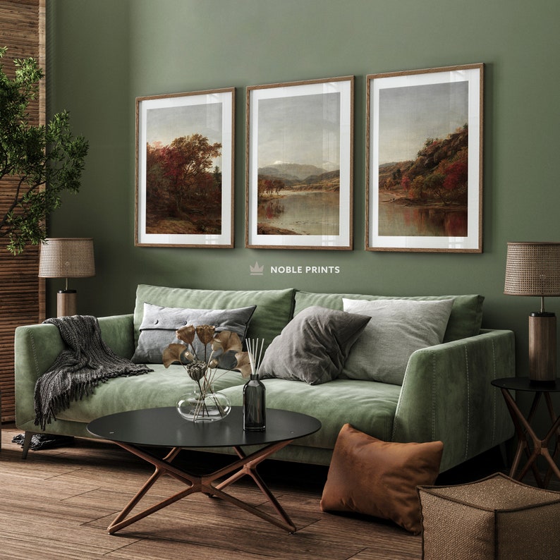 May include: Three framed landscape paintings of lakes and trees in a living room setting with a green sofa, a coffee table, and a throw pillow. The paintings are in a rustic style with muted colors.