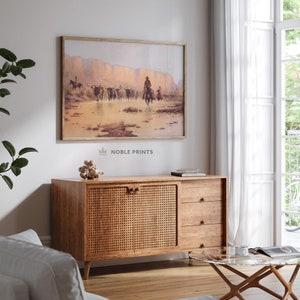 May include: A framed print of a vintage Western scene depicting cowboys on horseback herding cattle through a desert landscape. The print is in a wooden frame and hangs on a white wall above a wooden cabinet.