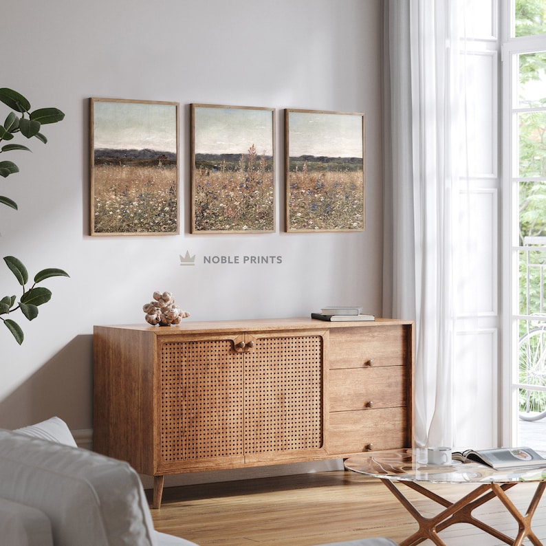 May include: Three framed landscape paintings with a muted color palette depicting a field of wildflowers. The paintings are hung on a white wall above a wooden cabinet with woven rattan doors.