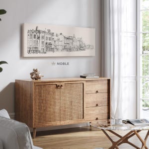 Puede incluir: Un aparador de madera con un panel de puerta tejido y tres cajones. Encima cuelga un boceto en blanco y negro de un paisaje urbano. La palabra "NOBLE" está impresa debajo de la obra de arte. También se ven una mesa de centro con tapa de cristal y una cortina blanca.