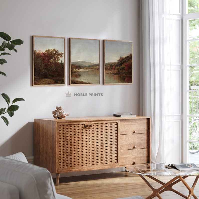 May include: Three framed landscape paintings with a muted color palette depicting scenes of nature with trees, water, and mountains. The paintings are hung on a white wall above a wooden cabinet with woven rattan doors.