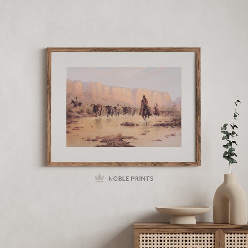 May include: A framed print of a painting depicting a cowboy riding a horse through a desert landscape with a herd of cattle. The scene is set against a backdrop of red rock cliffs and a clear blue sky.