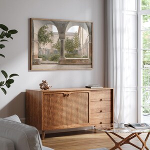 May include: A wooden sideboard with a woven rattan front, three drawers, and brass hardware. The sideboard is in front of a framed print of an arched courtyard with trees and stone columns.