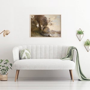 Puede incluir: Una pintura de paisaje enmarcada con un marco de madera clara cuelga sobre un sof&aacute; blanco. La pintura representa &aacute;rboles cerca de un cuerpo de agua, con tonos apagados de marr&oacute;n, verde y azul. Una manta verde y un coj&iacute;n redondo con un estampado de hojas est&aacute;n en el sof&aacute;.