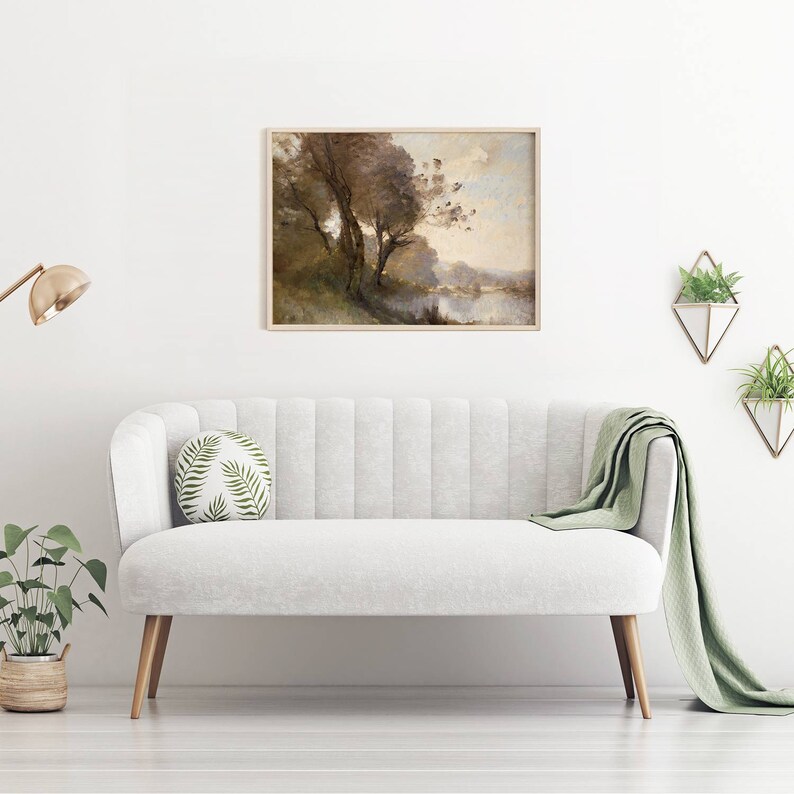 Puede incluir: Una pintura de paisaje enmarcada con un marco de madera clara cuelga sobre un sof&aacute; blanco. La pintura representa &aacute;rboles cerca de un cuerpo de agua, con tonos apagados de marr&oacute;n, verde y azul. Una manta verde y un coj&iacute;n redondo con un estampado de hojas est&aacute;n en el sof&aacute;.
