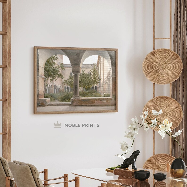 May include: A framed print of a watercolor painting depicting a courtyard with arched walkways and lush greenery. The painting is in a light brown frame and hangs on a white wall.