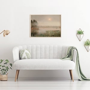 May include: A framed watercolor painting of a misty lake scene with trees on the shore and a sun shining through the fog. The painting is hanging on a white wall above a white sofa with a green throw blanket.