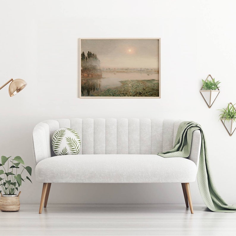 May include: A framed watercolor painting of a misty lake scene with trees on the shore and a sun shining through the fog. The painting is hanging on a white wall above a white sofa with a green throw blanket.