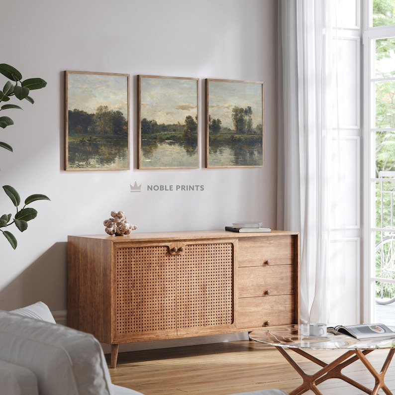 May include: Three framed landscape paintings with a muted color palette depicting a lake scene with trees and a cloudy sky. The paintings are hung on a white wall above a wooden cabinet with rattan doors.