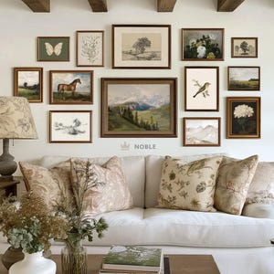 May include: A white sofa with floral patterned throw pillows sits in a living room with a gallery wall of framed artwork featuring nature scenes, landscapes, and botanical illustrations. The wall also includes a framed print with the word "NOBLE" in a crown.