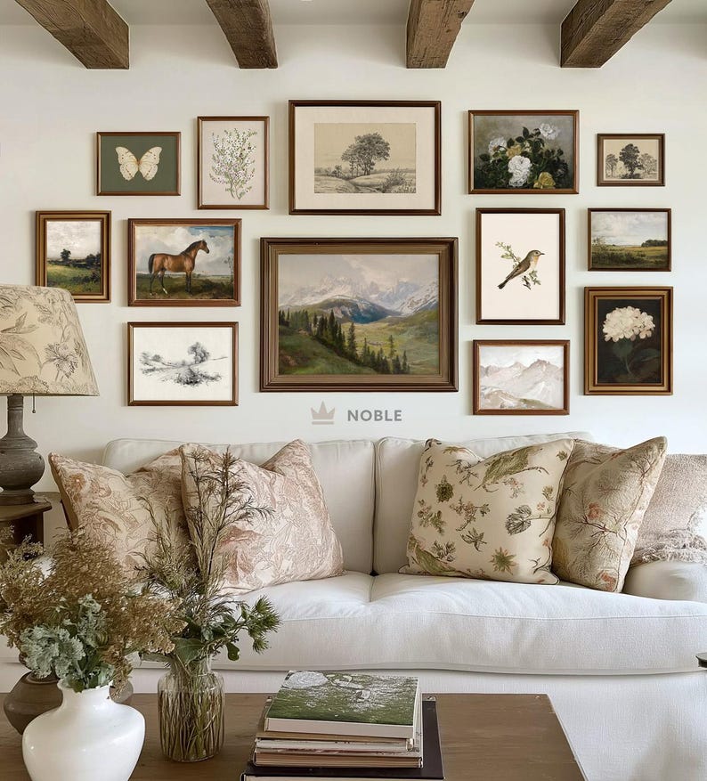 May include: A white sofa with floral patterned throw pillows sits in a living room with a gallery wall of framed artwork featuring nature scenes, landscapes, and botanical illustrations. The wall also includes a framed print with the word "NOBLE" in a crown.
