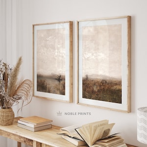 May include: Two framed landscape paintings with a muted brown and beige color scheme. The paintings are hung on a white wall above a wooden table with books and a vase of dried flowers.