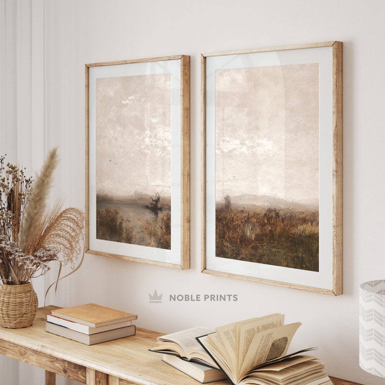 May include: Two framed landscape paintings with a muted brown and beige color scheme. The paintings are hung on a white wall above a wooden table with books and a vase of dried flowers.