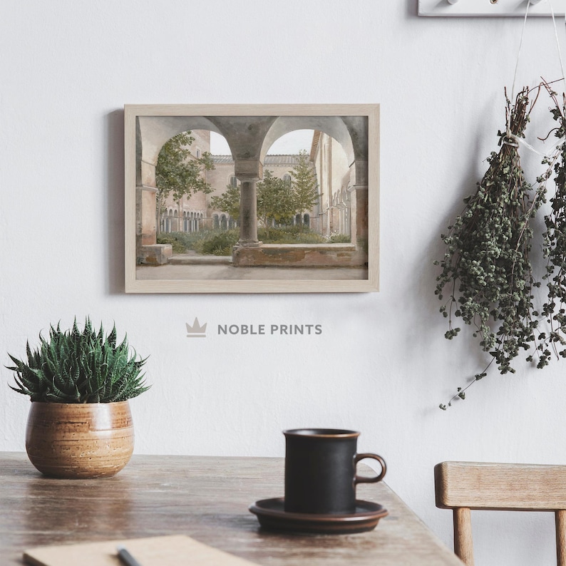 May include: A framed photograph of a courtyard with arched walkways and lush greenery. The courtyard is surrounded by stone walls and buildings. The photograph is in a light wood frame.