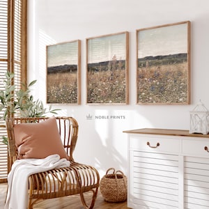 May include: Three framed landscape paintings with a muted color palette depicting a field of wildflowers. The paintings are hung on a white wall in a room with a wicker chair, a woven basket, and a white cabinet.