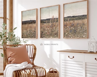 May include: Three framed landscape paintings with a muted color palette depicting a field of wildflowers. The paintings are hung on a white wall in a room with a wicker chair, a woven basket, and a white cabinet.