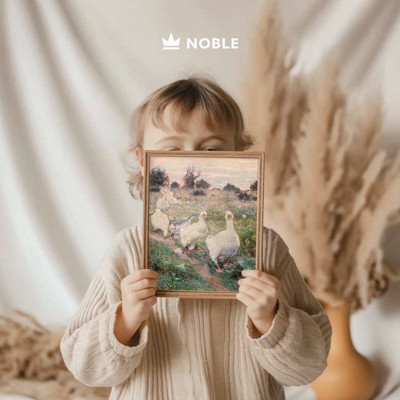 May include: A child holds a framed painting of white geese in a field. The painting is in a wooden frame with a light brown finish. The child is wearing a light brown sweater.