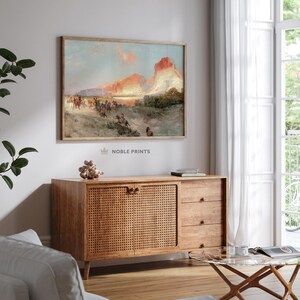 Puede incluir: Una pintura vintage que representa a un grupo de personas a caballo cabalgando por un paisaje des&eacute;rtico. La pintura presenta un cielo dram&aacute;tico con una puesta de sol naranja brillante y una gran formaci&oacute;n rocosa en el fondo. La pintura est&aacute; enmarcada en un marco de madera simple.