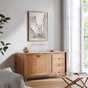 May include: A black and white sketch of a city street with tall buildings. The image is framed and hanging on a white wall above a wooden cabinet with woven rattan doors and drawers. The cabinet is in a living room setting with a glass coffee table and a potted plant.