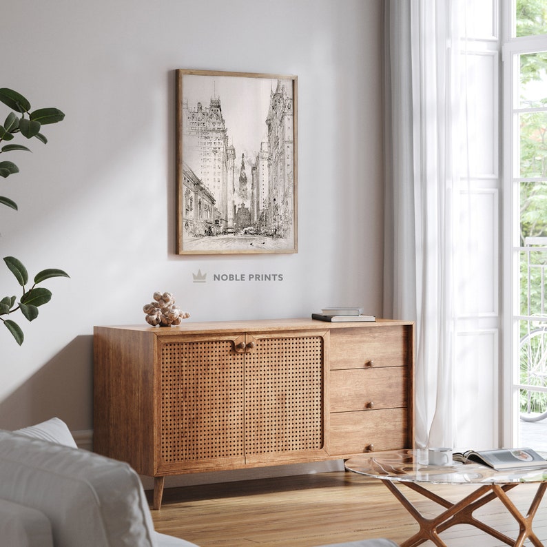 May include: A black and white sketch of a city street with tall buildings. The image is framed and hanging on a white wall above a wooden cabinet with woven rattan doors and drawers. The cabinet is in a living room setting with a glass coffee table and a potted plant.