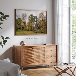Op de afbeelding: Een ingelijste foto van een boslandschap met bomen in verschillende tinten groen en geel. De bomen bevinden zich op de achtergrond en de voorgrond is een grasveld. De foto hangt aan een witte muur.