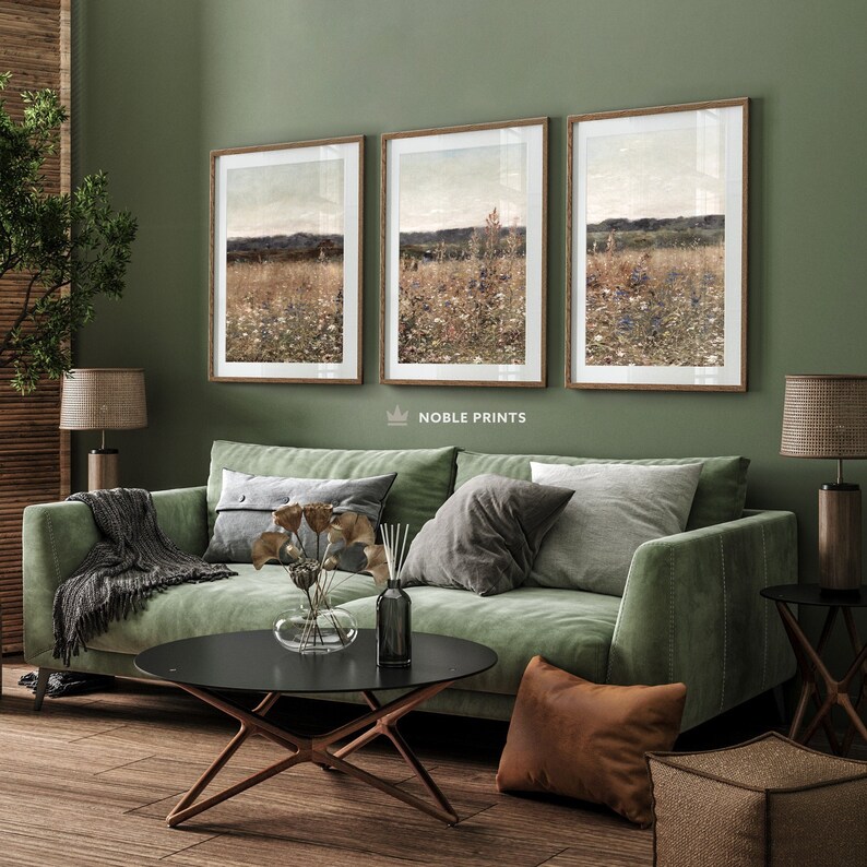 May include: Three framed landscape paintings of a field of wildflowers with a distant horizon. The paintings are hung on a sage green wall above a green velvet sofa with brown throw pillows.