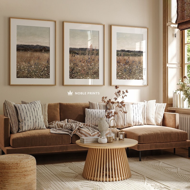 May include: A brown sectional sofa with a wooden coffee table and a woven ottoman in front of it. The sofa has a variety of throw pillows and a blanket. Three framed landscape prints hang on the wall behind the sofa. The text "NOBLE PRINTS" is printed below the prints.