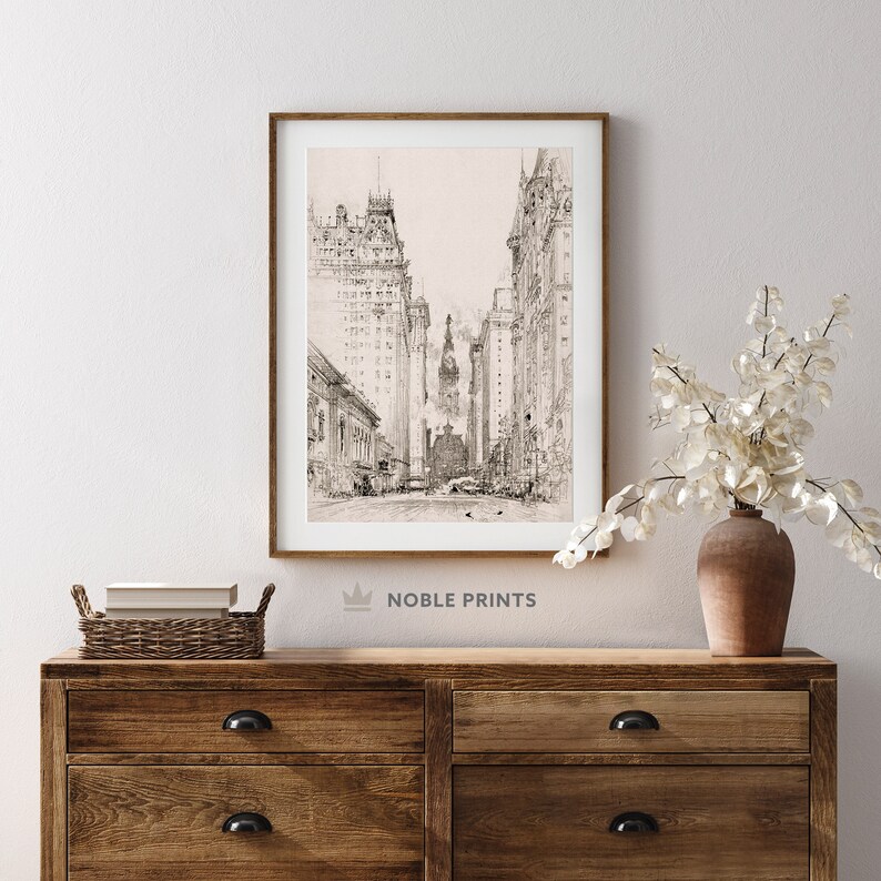 May include: A black and white line drawing of a city street with tall buildings on either side. The drawing is framed in a light wood frame and is hanging on a white wall.