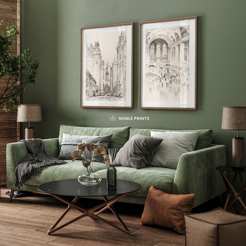 May include: Two black and white framed illustrations of cityscapes hang on a sage green wall above a green velvet sofa. The sofa has a black coffee table in front of it and a brown throw pillow on the floor.