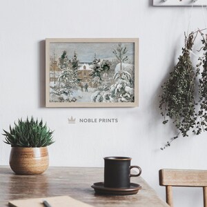 May include: A framed winter landscape painting with snow-covered trees and a building. The artwork is in a light wooden frame and hangs on a white wall. A small potted plant and a black mug on a saucer sit on a wooden table.