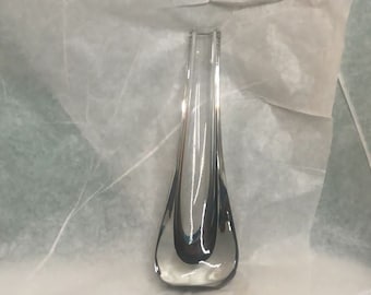 May include: A clear glass vase with a teardrop-shaped body and a narrow, rectangular opening. The vase reflects the surrounding light, creating highlights and shadows. The vase is displayed on a white surface.