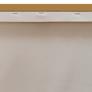 May include: A close-up of the back of a stretched canvas painting. The canvas is white and is stretched over a wooden frame. The frame is painted a light brown color. The canvas is secured to the frame with staples.