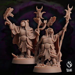 May include: Two painted miniature figures of anthropomorphic badger-like creatures wearing robes and holding staffs. The figures are standing on circular bases with a textured surface. The figures are painted in shades of brown and have detailed features.