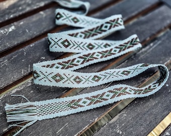 Può includere: Una lunga cintura in tessuto intrecciato con una base azzurro chiaro e un motivo geometrico ripetitivo in marrone scuro e verde bosco. La cintura presenta una trama piatta e testurizzata e termina con fili sciolti che formano una frangia.