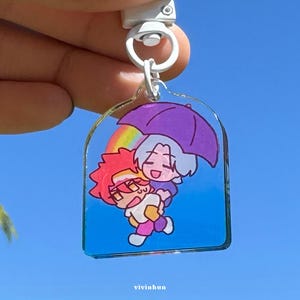 Könnte beinhalten: Ein klarer Acryl-Schlüsselanhänger mit einer Cartoon-Illustration von zwei Figuren unter einem lila Regenschirm, mit einem Regenbogen im Hintergrund. Der Schlüsselanhänger hat einen silberfarbenen Verschluss und einen blauen Hintergrund. Der Text "vivinhun" befindet sich unten.
