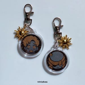May include: Two clear acrylic keychains with silver-colored clasps. Each keychain features a circular design with a cartoon face and a gold-colored sun charm. The keychains are on a light blue background.