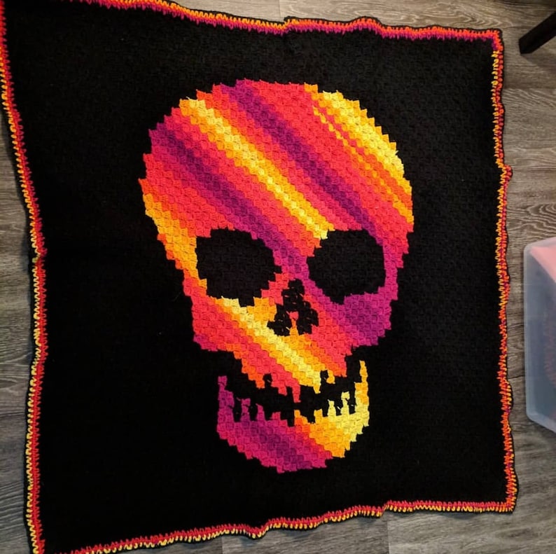 Może przedstawiać: Czarny koc szydełkowy z kolorowym wzorem czaszki. Czaszka jest wykonana z pomarańczowej, ż&oacute;łtej, czerwonej i fioletowej przędzy. Koc ma czarną obw&oacute;dkę z czerwoną i pomarańczową lam&oacute;wką.