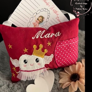 Könnte beinhalten: Ein rosa Zahnfee-Kissen mit einem weißen Zahn, der darauf gestickt ist. Der Zahn hat eine goldene Krone und Flügel. Das Kissen hat eine Tasche an der Seite und ist mit dem Namen "Mara" bestickt.