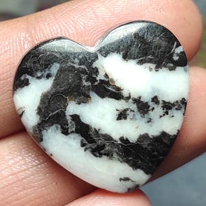 Puede incluir: Una piedra en forma de corazón con un llamativo patrón en blanco y negro. La superficie pulida revela un efecto veteado, con vetas negras que recorren la base blanca. La piedra se sostiene en una mano, mostrando su tamaño y forma.