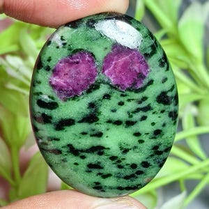 Puede incluir: Una piedra preciosa pulida de forma ovalada que presenta una base de color verde vibrante con motas negras dispersas y dos inclusiones prominentes e irregulares de color magenta. La superficie es lisa y reflectante, mostrando un patrón mineral natural.